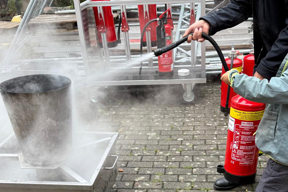 Erwachsender sprüht einen Wasserstrahl aus einem Feuerlöscher auf einen Brandsimulator. Feuer ist aus.